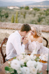 Wedding at an old winery villa in Tuscany, Italy. The wedding couple is sitting at the dinner table on the roof of an old villa, the groom holds the bride by the hands.