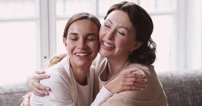 Happy laughing affectionate beautiful middle aged mother cuddling, feeling proud of sincerely smiling young mixed race grown daughter, enjoying sweet tender family moment together in living room.