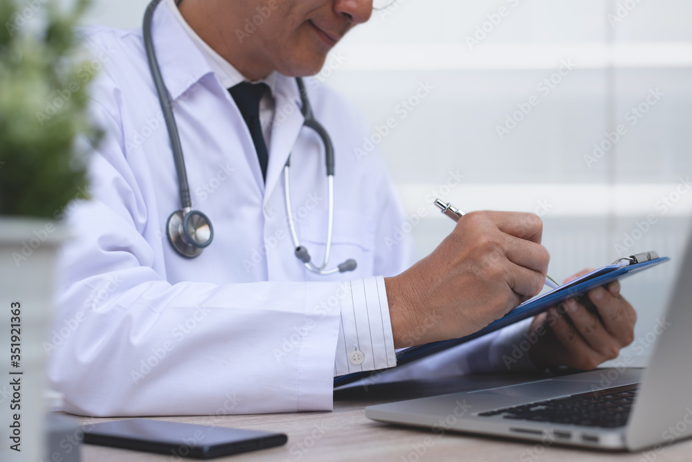 Sticker Asian doctor recording patient information or writing prescription document on clipboard while working on laptop computer in medical office