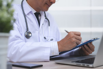 Asian doctor recording patient information or writing prescription document on clipboard while working on laptop computer in medical office