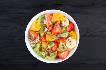 Fruit salad with chia seeds in the white  bowl in the center of  the black wooden background. Top view. Closeup.