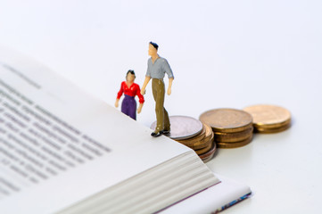Miniature couple walking up stacked coins onto a book in a concept of financial barriers to higher education, study and career over a white background with copy space