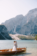 Fototapeta na wymiar Bride and groom sailing in wooden boat, with oars at Lago di Braies lake in Italy. Wedding in Europe - Newlyweds are standing embracing in a boat.