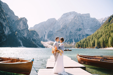 The bride and groom walk along a wooden boat dock at the Lago di Braies in Italy. Wedding in Europe, on Braies lake. Newlyweds walk, kiss, hug on a background of rocky mountains.