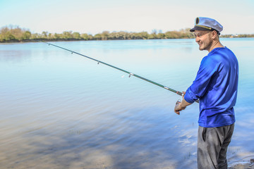 a fisherman in a blue T-shirt and white cap is standing on the river with a fishing rod