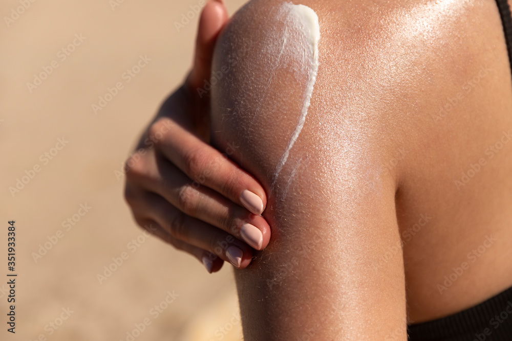 Wall mural Young woman applying sun cream or sunscreen on her tanned shoulder to protect her skin from the sun. Shot on a sunny day with blurry sand in the background