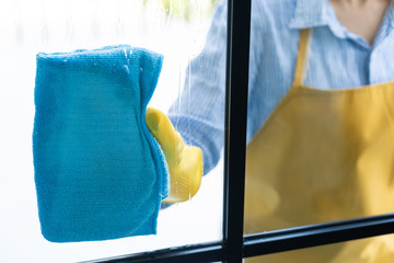Young asian Woman in bright yellow gloves Washing or Cleaning Window  the glass with blue rag wipe and detergent of her apartment from dust and stains.