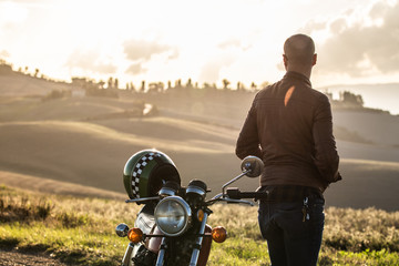 Man standing next his motorbike taking along a country road on hills landscape at sunset. Tuscany,...