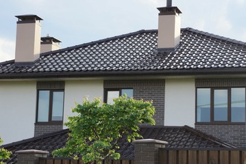 part of a private brick house with windows under a brown tiled roof with a chimney and green decorative tree