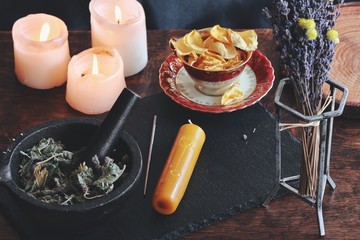 Symbols and sigils carved onto a yellow gold color candlestick, that is laying on a black surface. Witchy elements in a slightly blurred background, like burning candles and dried flowers