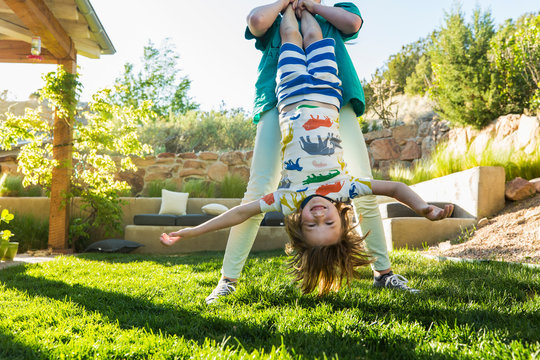 teenager sister holding her younger brother upside down on green lawn at sunset