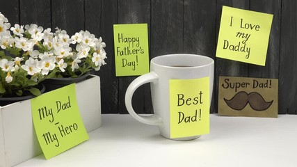 Cup of hot tea with greeting sticker in honor of Father's Day is standing near primrose flowers on a white table against the background of a wooden background