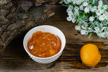Apricot and peach jam in a small deep dish, sweet orange jam for dessert. Next to a whole fruit, apricots are marinated in syrup. Old wooden background and snags with bark with white flowers