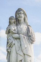 Fototapeta na wymiar Very ancient stone statue of the Virgin Mary with the baby Jesus Christ against blue sky. Religion, faith, eternal life, God, the soul concept.