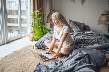 Happy beautiful blonde woman working, making video call, watching movie on a laptop in cozy home bedroom modern interior. Home quarantine Covid-19 pandemic Corona virus. Remote work from home concept.