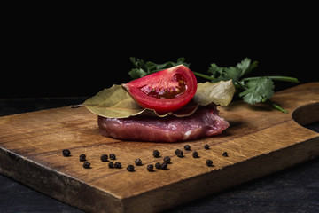 Cooking concept. one piece of meat (pork) is laying on a wooden board. On top lies a bay leaf and a slice of tomato. Around the meat are spices and cilantro. photo on a black background.
