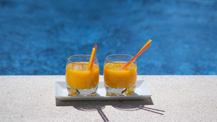 Two glasses with tropical, mango, orange juice with ice cubes on swimming pool or sea blurred background. Exotic alcoholic cocktail with straw on a sunny day near the pool. Luxury drink.