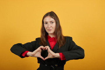 Young Attractive Brunette Woman In Black Stylish Suit, Red Shirt On Yellow Background, Happy Female Shows Hands Heart Shape. The Concept Fell In Love