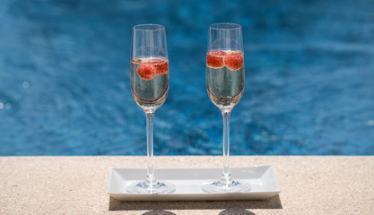 Close up of glasses with champagne or prosecco with raspberry on swimming pool or sea blurred background. Romantic weekend, vacation for two. Luxury drink.