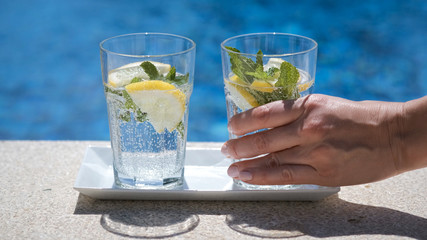 Hand of a young woman takes a glass of fresh lemon infused mineral water with gas and mint. Pouring sparkling water with bubbles in sunny day on blurred pool or sea background. Refreshing beverages.