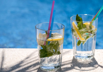 Fresh lemon infused mineral water with gas or soda drinks in glasses with mint in the shadow of palm leaves. Pouring sparkling water with bubbles in sunny day with on blurred pool or sea background.
