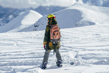 Girl with bright helmet and colourful backpack on a snowboard admires snow-capped mountain peaks. She is choosing the further line for freeride.