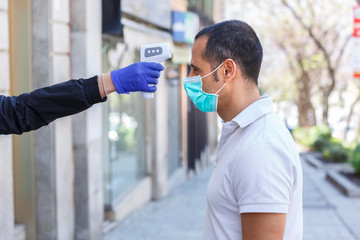 A uomo moro con mascherina chirurgica viene presa la temperatura del corpo con termometro elettronico digitale, isolato su sfondo urbano