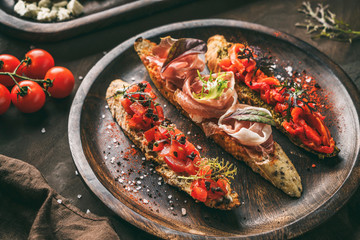 Variety of bruschetta with prosciutto, tomatoes, parmesan cheese, sauce pesto, grilled pepper on ciabatta bread on wooden plate over dark background. Sandwiches set of wine, top view