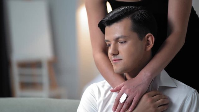 The Young Handsome Man Sits On The Sofa In The Stylish Bedroom .The Woman Wearing Black Dress Is Standing Behind Him And Unbuttoning His Shirt. The Man Is Smiling.