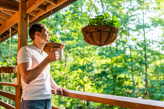 Hanging potted plant flowerpot with man drinking coffee tea mug cup on porch of log cabin cottage house with green color