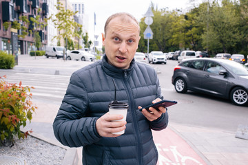 Obraz premium A caucasian positive surprised man in a gray jacket with a white paper cup of Latte coffee or tea with lid and straw and phone on his hand on the city background. Eco. break. Close up