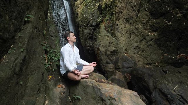 handsome young man with short dark haircut closes eyes and meditates sitting on rock in yoga lotus position against waterfall in national park