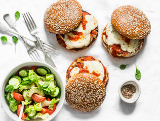 Served appetizers table - homemade whole grain bun, grilled chicken patty, tomato sauce, mozzarella cheese burgers and tomato, cucumber, avocado salad on a light background, top view