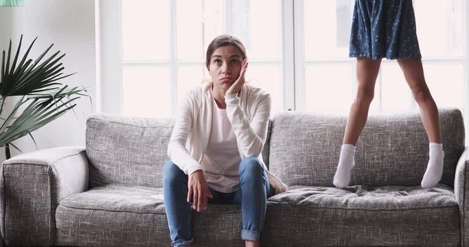 Desperate young mixed race mother sitting on sofa, feeling annoyed of naughty small daughter at home. Stressed nanny babysitter irritated by child girl bad behavior, disrespectful relations concept.