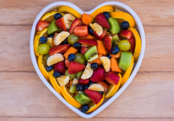 Mixed colorful fruit salad in a white ceramic plate heart shaped. Wooden background