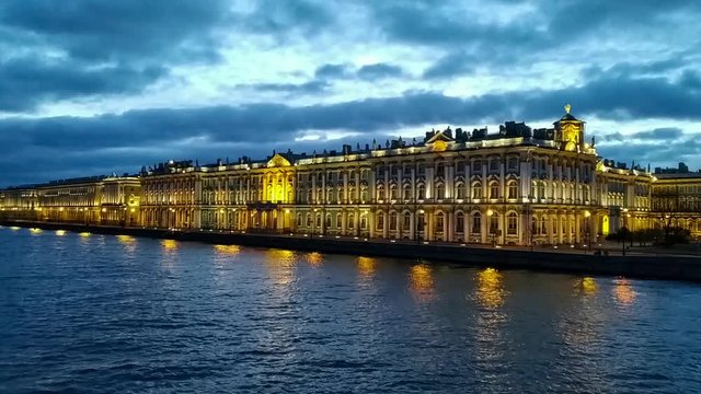 Cityscape of night Saint Petersburg just before sunrise. The building of the Winter Palace is illuminated by lights. Small waves on the Neva river. Few cars drives along the road. 