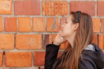 Fototapeta na wymiar Smiling girl in sunglasses and black raincoat on a red brick wall background talking on the phone. Beautiful young blonde long hair hipster woman on the street with smartphone in hands,lifestyle photo