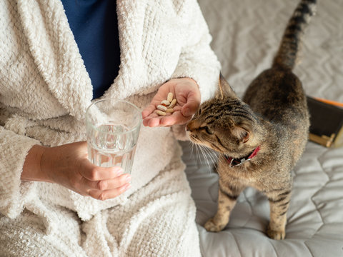 Elderly Woman In  White Terry Cloth Robe Sitting On The Bed And Washes Down The Pills With A Glass Of Water