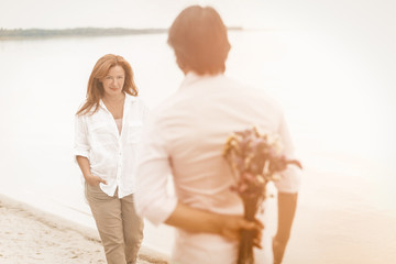 Romantic date on seashore. Focus on woman in a white shirt. Rear view of man holding a bouquet of flowers behind his back in the foreground. Toned image.