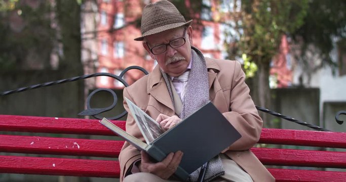 Lonely senior man flipping photo album on a bench in park square