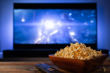 A wooden bowl of popcorn and remote control in the background the TV works. Evening cozy watching a...