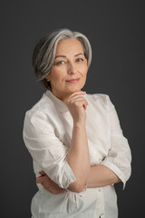 Beautiful gray-haired woman gently smiles while touching her chin with hand and looking at camera. Intelligent beautiful mature lady wearing white shirt standing alone on grey background