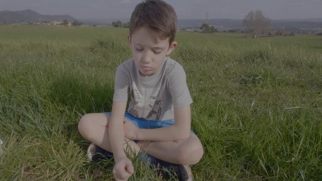 Sad Spanish Child Sits Down while Pulls Up Grass, Outdoors Daylight, Spain, 4K