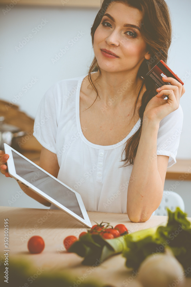 Wall mural Young woman looking for a new recipe for cooking in a kitchen. Housewife is making online shopping by tablet computer and credit card