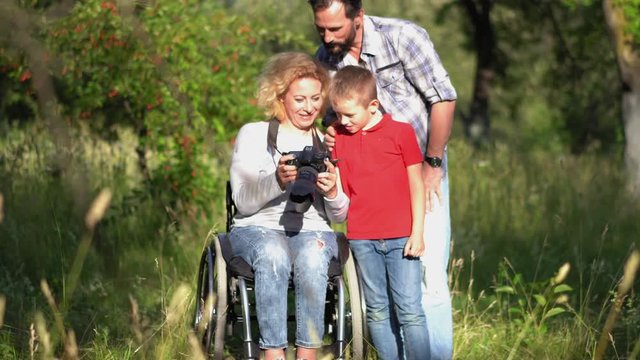 Woman photographer in a wheel chair taking a photos of father with daughter outdoors in the park or backyard fruit tree garden. Family concept.