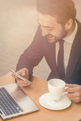 Happy businessman reading messages on mobile phone. Laughing man drinks coffee in street cafe and chatting on smartphone while working on computer. Freelance concept. Toned image