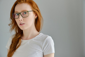 A red-haired girl in glasses and a white T-shirt stands on a gray background in a half-turn and looks surprised into the frame
