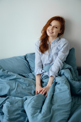 Obraz premium A beautiful red-haired girl in striped pajamas sits in a blue bed and smiles cutely. 