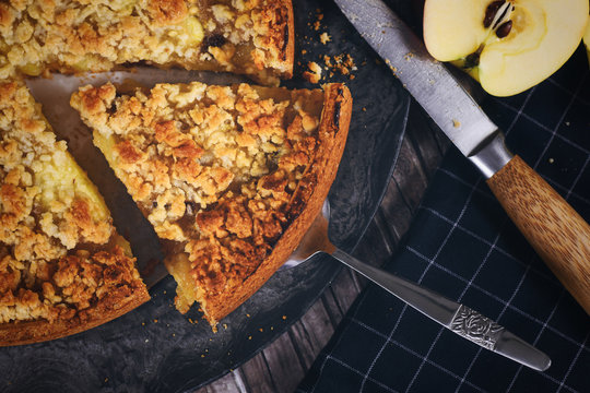 Slice of traditional European apple pie with topping crumbles on cake server surrounded by fresh appls and knife