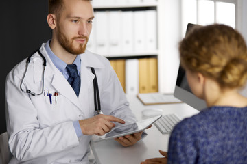 Fototapeta premium Friendly red-bearded doctor and patient woman discussing current health examination while sitting and using tablet computer in clinic. Medicine concept
